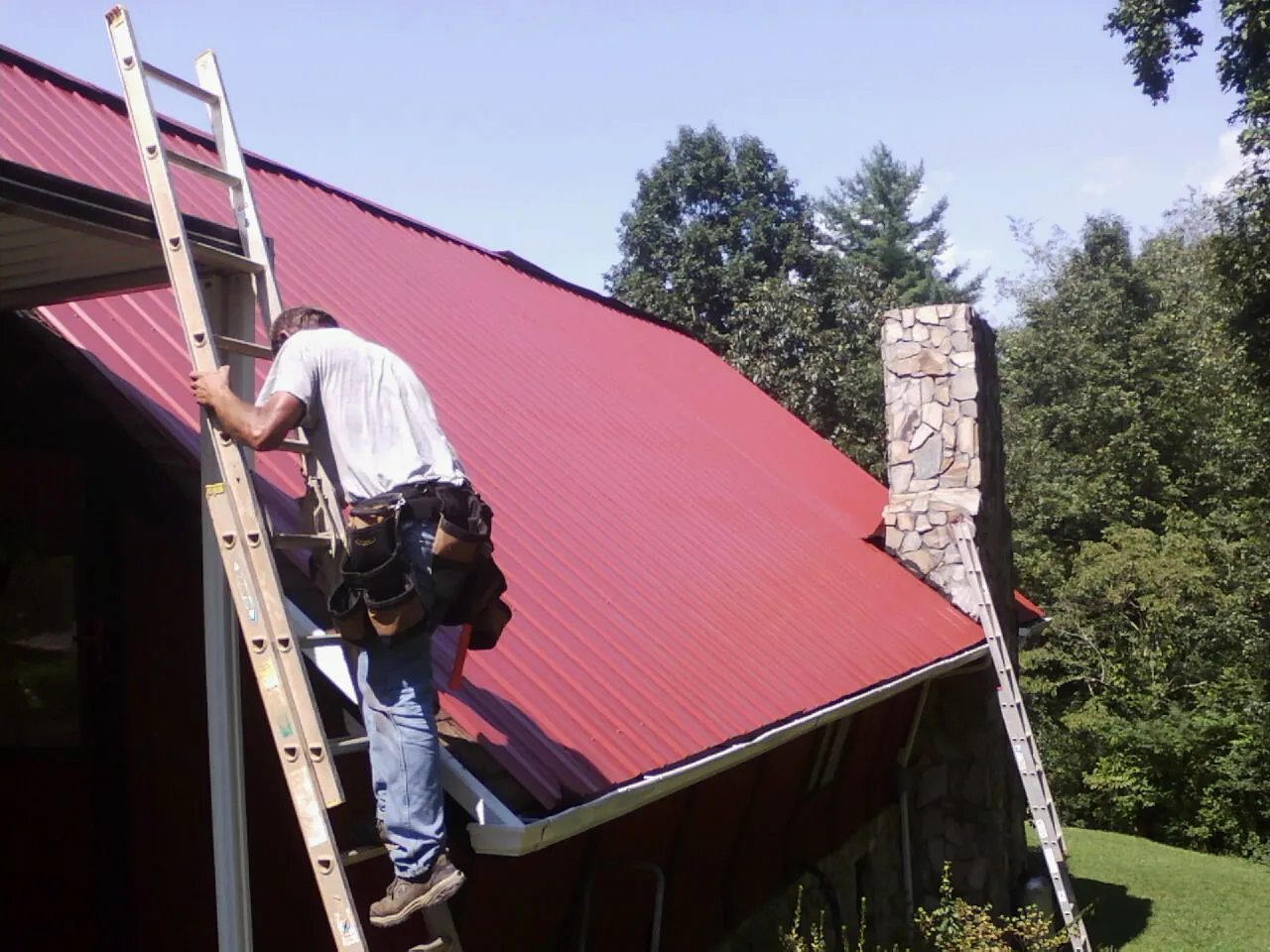 Roofer on a ladder beside a red metal roof and stone chimney — Soffit & Fascia Repair by Roofing Brothers in Haines
